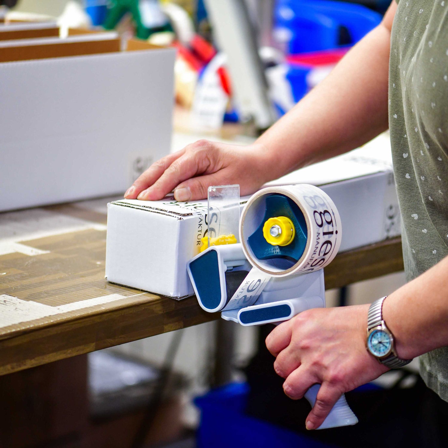 Ein Mitarbeiter in der Verpackungsabteilung verschließt einen weißen Karton mit einem Handabroller und Klebeband, das mit dem Logo „giese MANUFAKTUR“ bedruckt ist. Im Hintergrund sind weitere Verpackungsmaterialien und Kartons zu sehen.