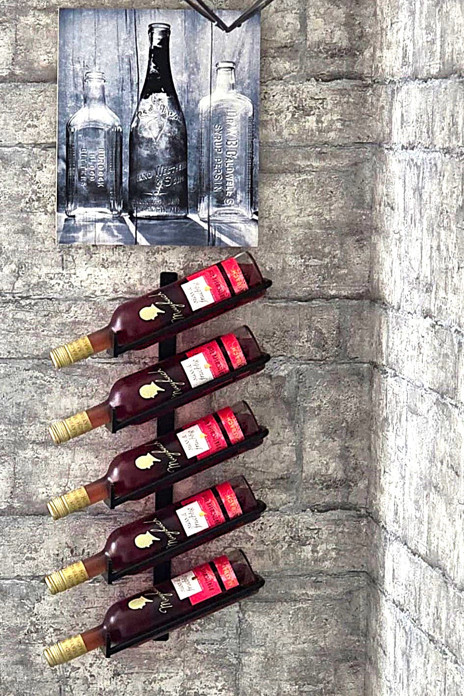 Hier sieht man ein schwarz, mattes Weinregal zur Wandmontage mit fünf Ablagen für Weinflaschen; auf dem Bild sind fünf Weinflaschen in den Halterungen eingelegt. Das Weinregal ist hier exe,plarisch an einer Wand montiert. Darüber hängt ein Bild mit Flaschen.