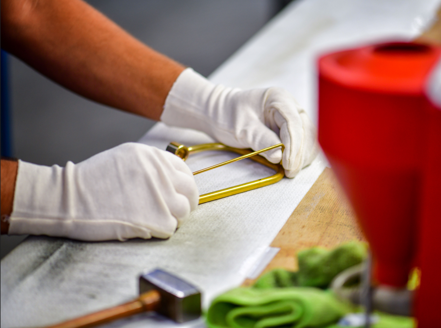 Zwei Hände mit weißen Handschuhen bearbeiten ein Bauteil aus Messing, vermutlich einen Dusch- oder Aufbewahrungskorb, auf einer Werkbank. Im Vordergrund sind ein Hammer und Reinigungstücher zu sehen – die Szene zeigt einen Produktionsschritt in der manuellen Fertigung.